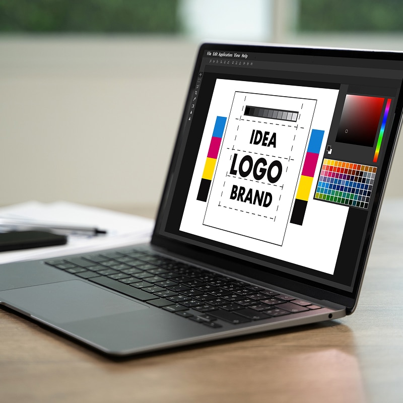 Ordinateur portable avec logiciel de design graphique. L'écran affiche "IDEA LOGO BRAND", des barres CMJN et une palette de couleurs.